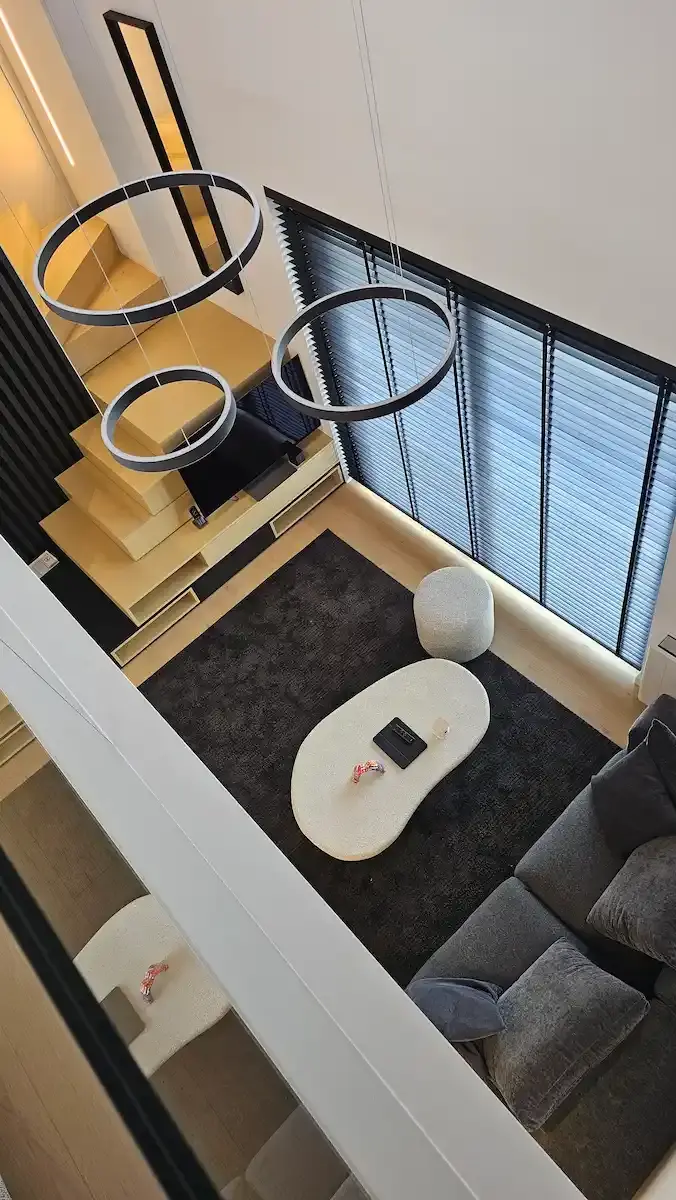 Modern living space with black kitchen counter, two black bar stools, gray sofa with pillows, light wood floor, and a colorful tattooed hands artwork on a white wall.
