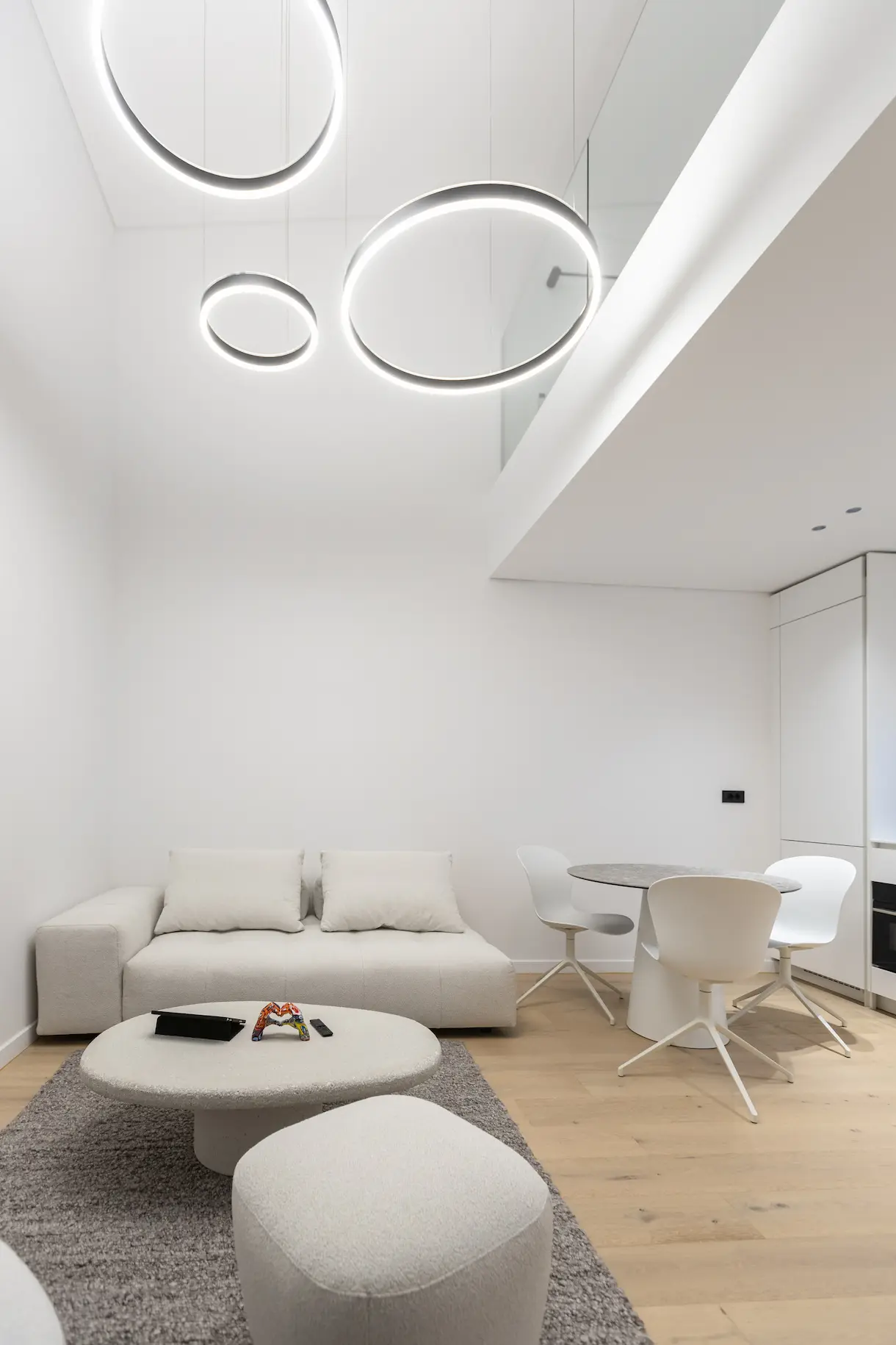 Minimalist living room with a cream sofa, round coffee table, ottoman, dining table with three white chairs, and modern circular ceiling lights.
