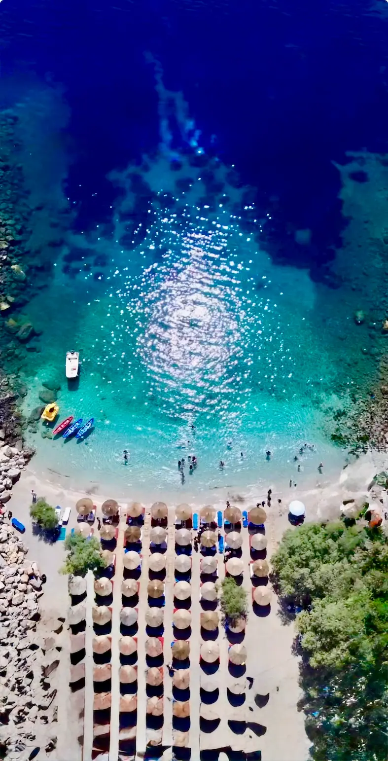 Aerial view of a beach with rows of umbrellas and sun loungers next to clear turquoise water where people swim and boats are anchored.
