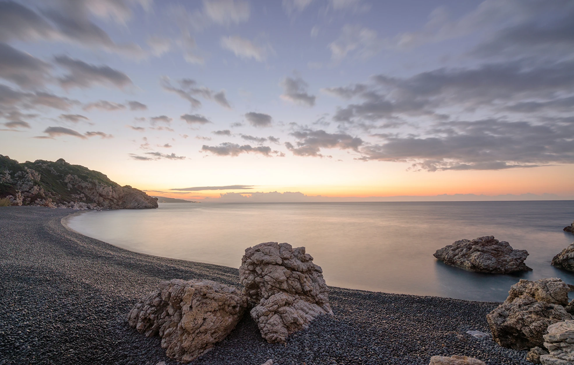 Pebble beach with large rocks and a calm sea at sunset under a partly cloudy sky.