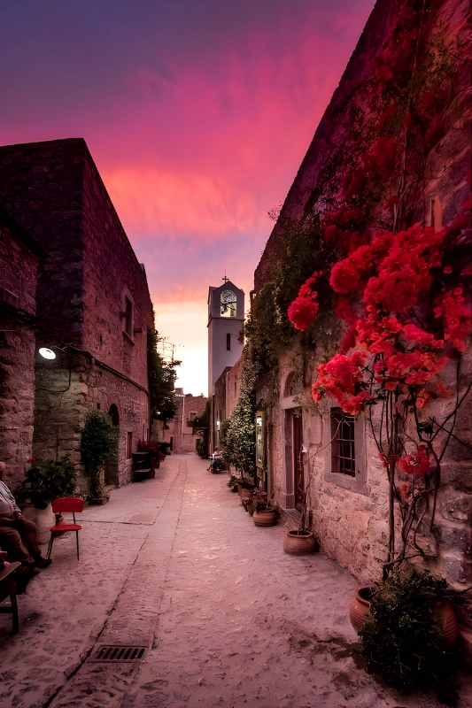 Narrow cobblestone street with stone buildings, vibrant red flowers climbing a wall, and a white church tower under a purple and orange sunset sky.