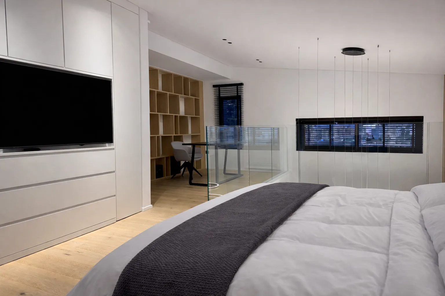 Modern bedroom with a large bed, gray throw blanket, wall-mounted TV, glass railing, and a study nook with shelves and a chair.