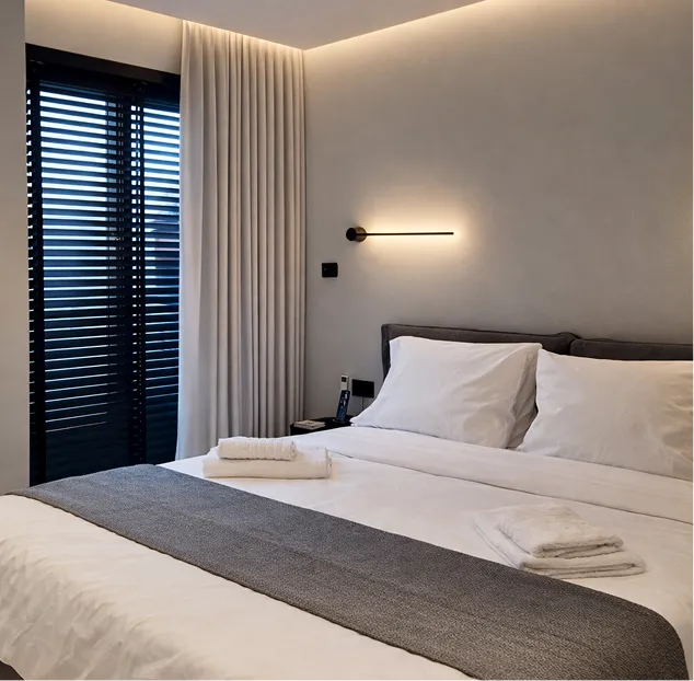 Modern bedroom with a large bed featuring white linens, gray blanket, folded towels, black nightstand, and wall-mounted linear lights.