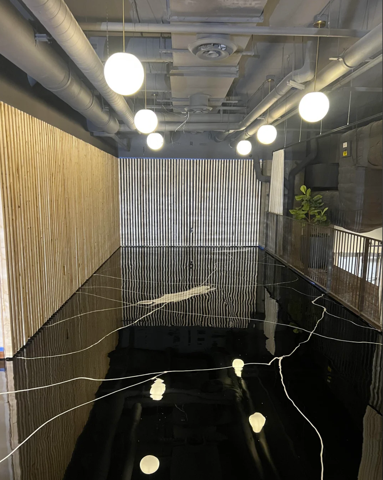 Indoor area with glossy black floor featuring white veining, wood-paneled wall on the left, striped wall at the end, metal railing on the right, ceiling with exposed ducts, and round hanging lights reflecting on the floor.