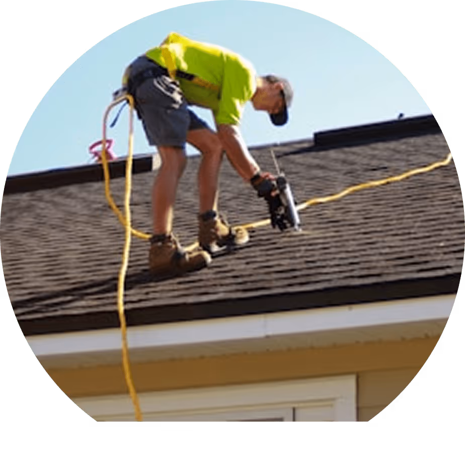 Ein Arbeiter in gelbem Hemd und blauer Kappe befestigt Schindeln auf einem Dach mit einem Nagelgerät, gesichert mit einem gelben Seil.