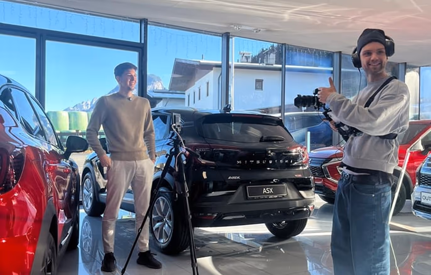 Zwei Männer in einem Autohaus: einer steht vor einem schwarzen Mitsubishi ASX, der andere filmt ihn mit Kamera und Daumen hoch.