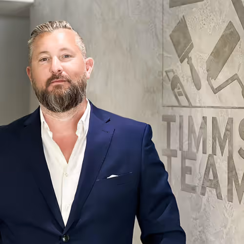 Mann mit kurzem grauem Haar und Bart in blauem Anzug vor einer Wand mit dem Schriftzug 'TIMMS TEAM'.