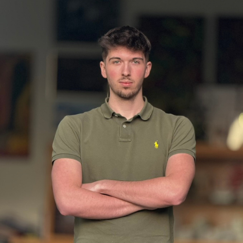 Junger Mann in einem weißen Pullover sitzt an einem Tisch und lächelt.