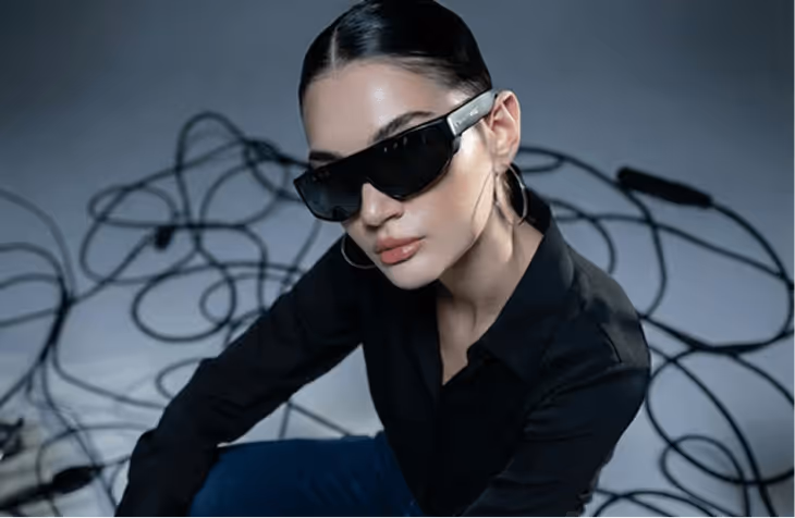 Woman with slicked-back hair wearing Mixverse black sunglasses and hoop earrings, dressed in a black shirt, sitting amid tangled black cables.