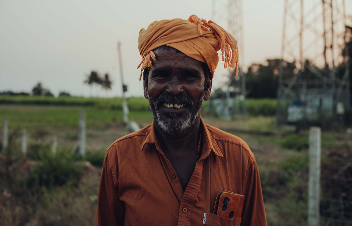 Lächelnder Mann mit orangefarbener Turban und Hemd vor ländlicher Kulisse bei Sonnenuntergang.