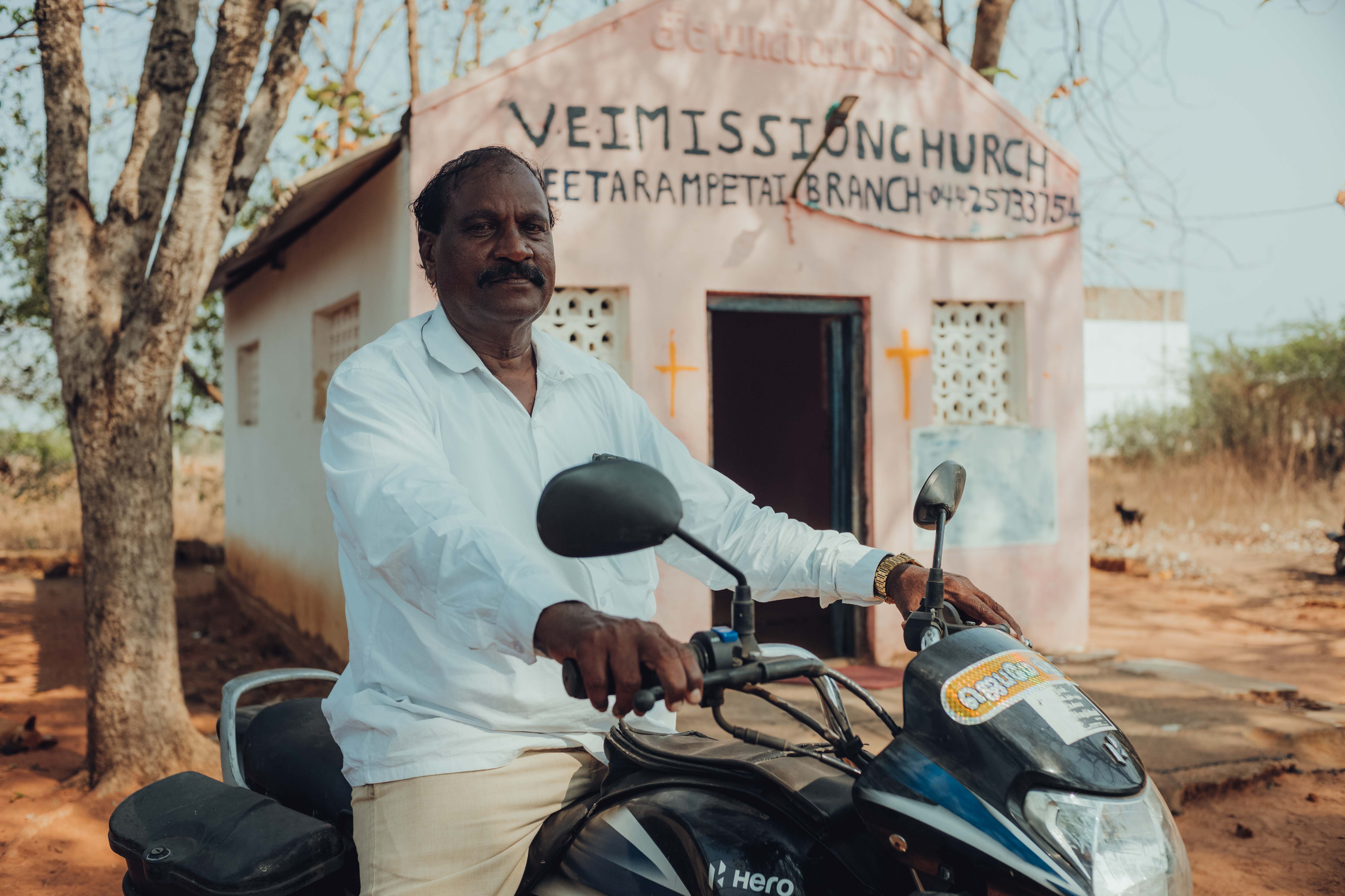 Man in white shirt sitting on a black Hero motorcycle in front of a small church building.