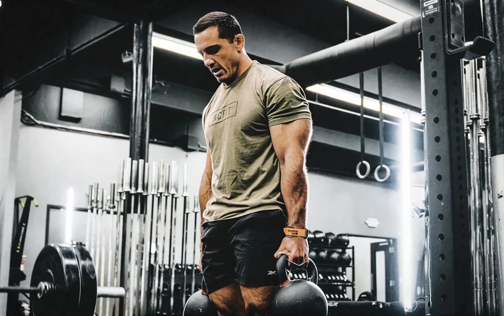 Man lifting two kettlebells in a gym with weightlifting equipment and gymnastic rings in the background.