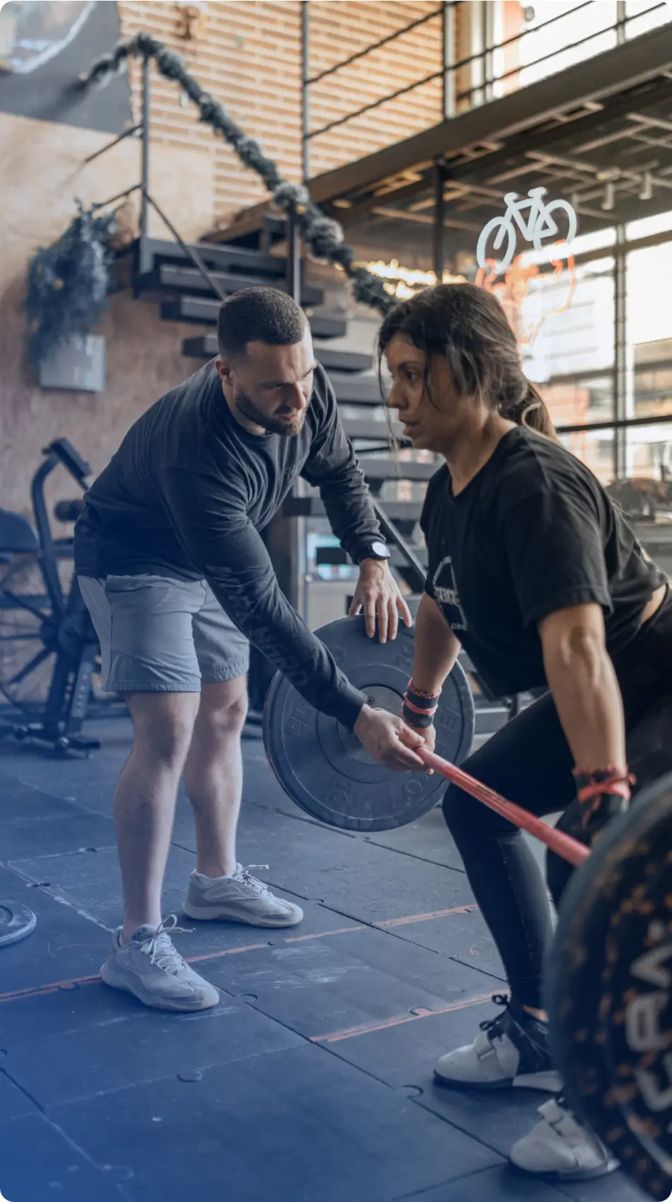 A male coach assists a woman lifting a barbell during a workout in a gym.
