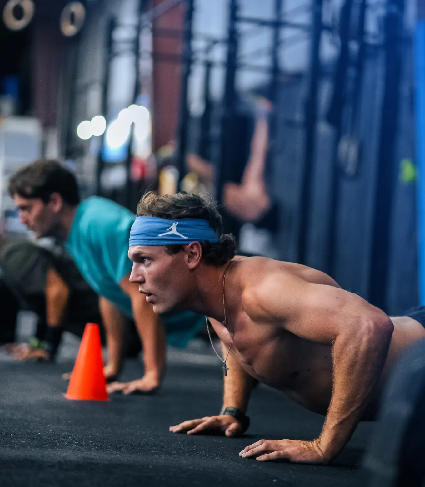 Shirtless man wearing a blue headband doing push-ups in a gym, with another person exercising in the background.