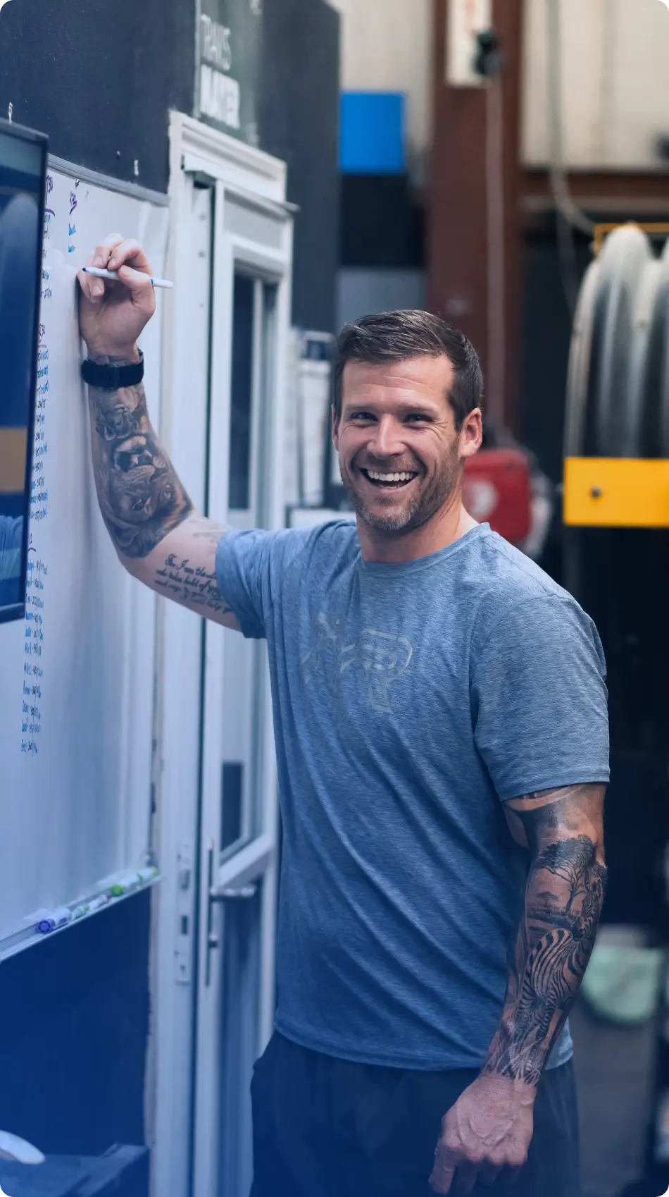 Smiling man with tattoos on both arms writing on a whiteboard in an indoor gym setting.