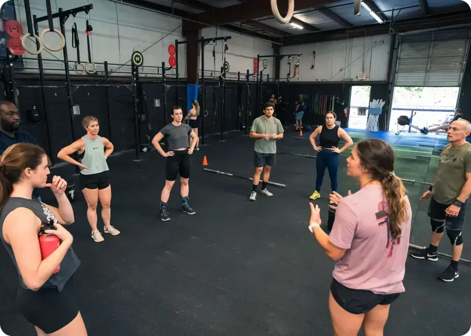 Group of people in athletic wear standing in a circle inside a gym listening to a woman speaking.