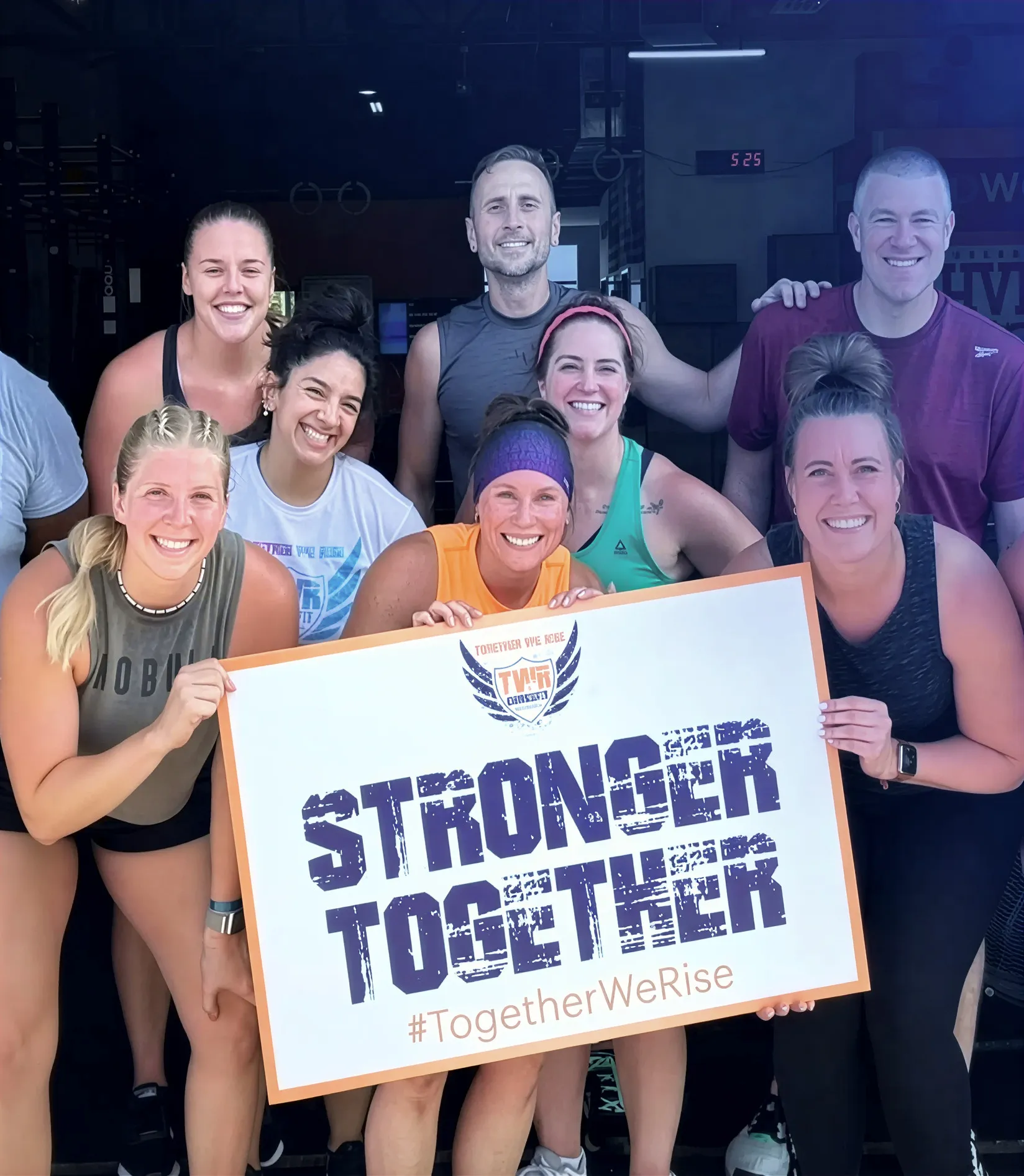 Group of smiling adults at a gym holding a sign that says 'STRONGER TOGETHER #TogetherWeRise'.