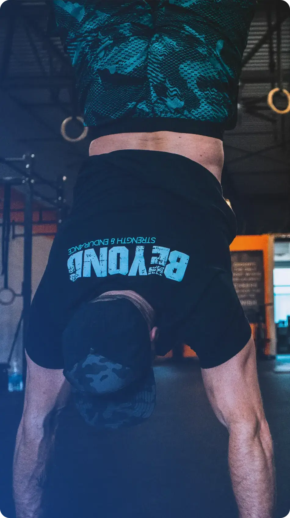 Man performing a handstand in a gym wearing a black shirt with 'BEYOND Strength & Endurance' text and a camo cap.