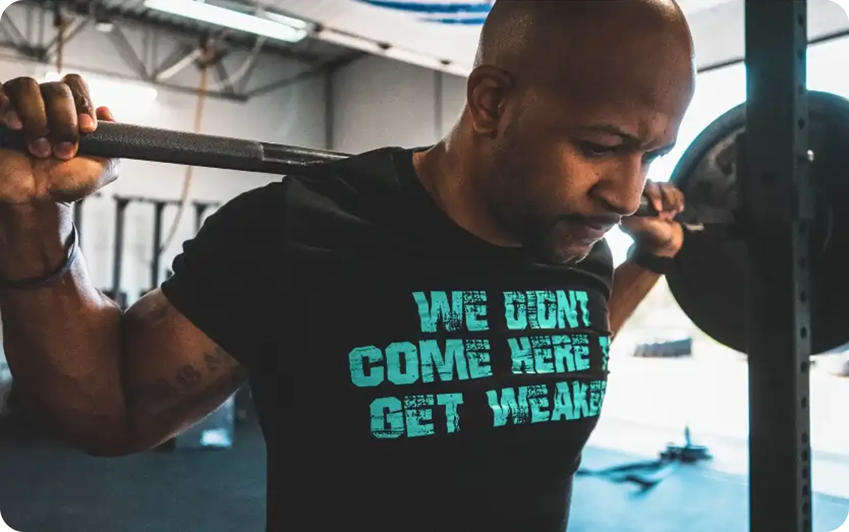 Man lifting a weighted barbell on his shoulders in a gym wearing a black shirt that reads 'We didn't come here to get weaker.'
