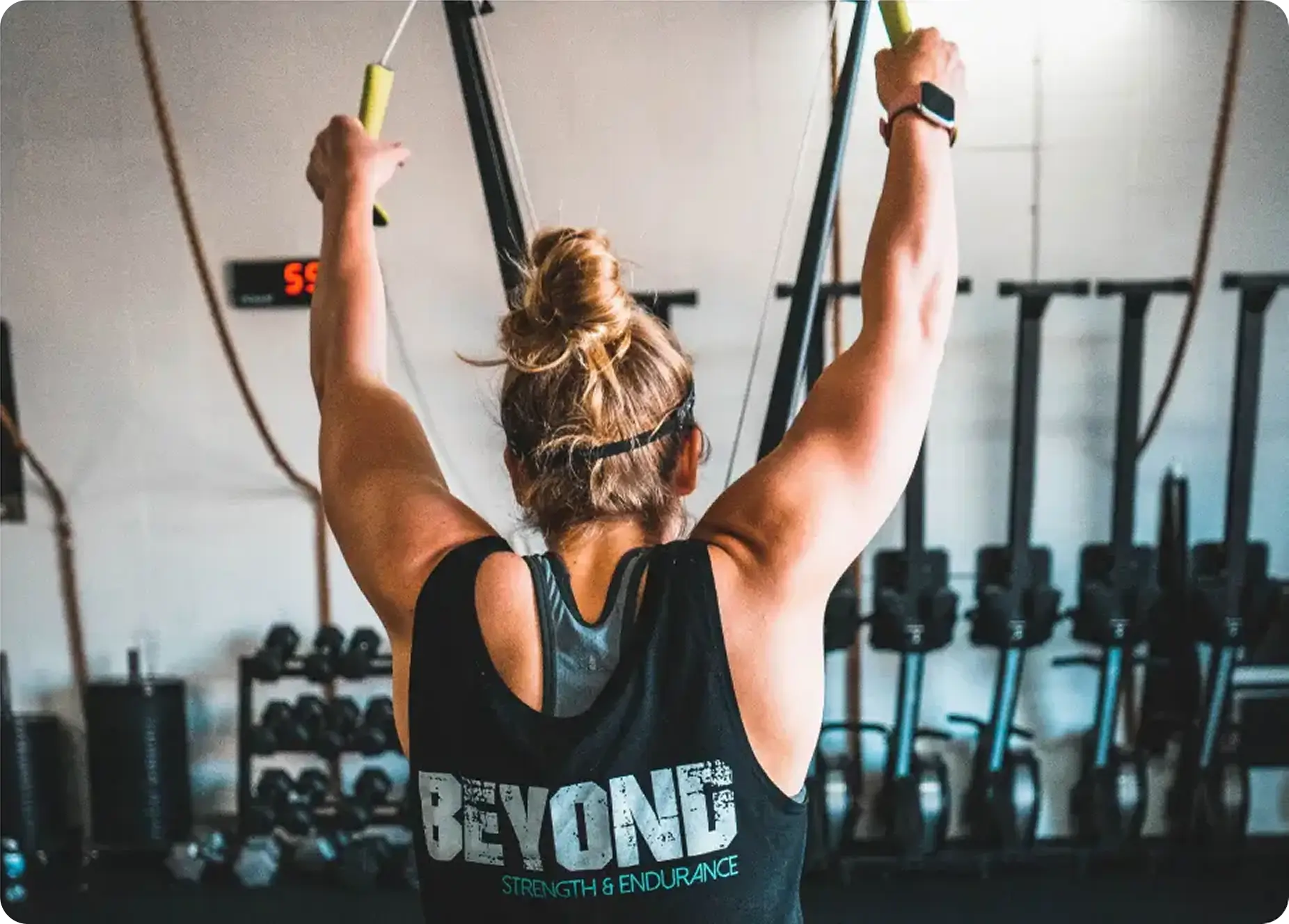 Woman with a bun hairstyle wearing a black tank top that says 'BEYOND Strength & Endurance' doing a lat pulldown exercise in a gym.