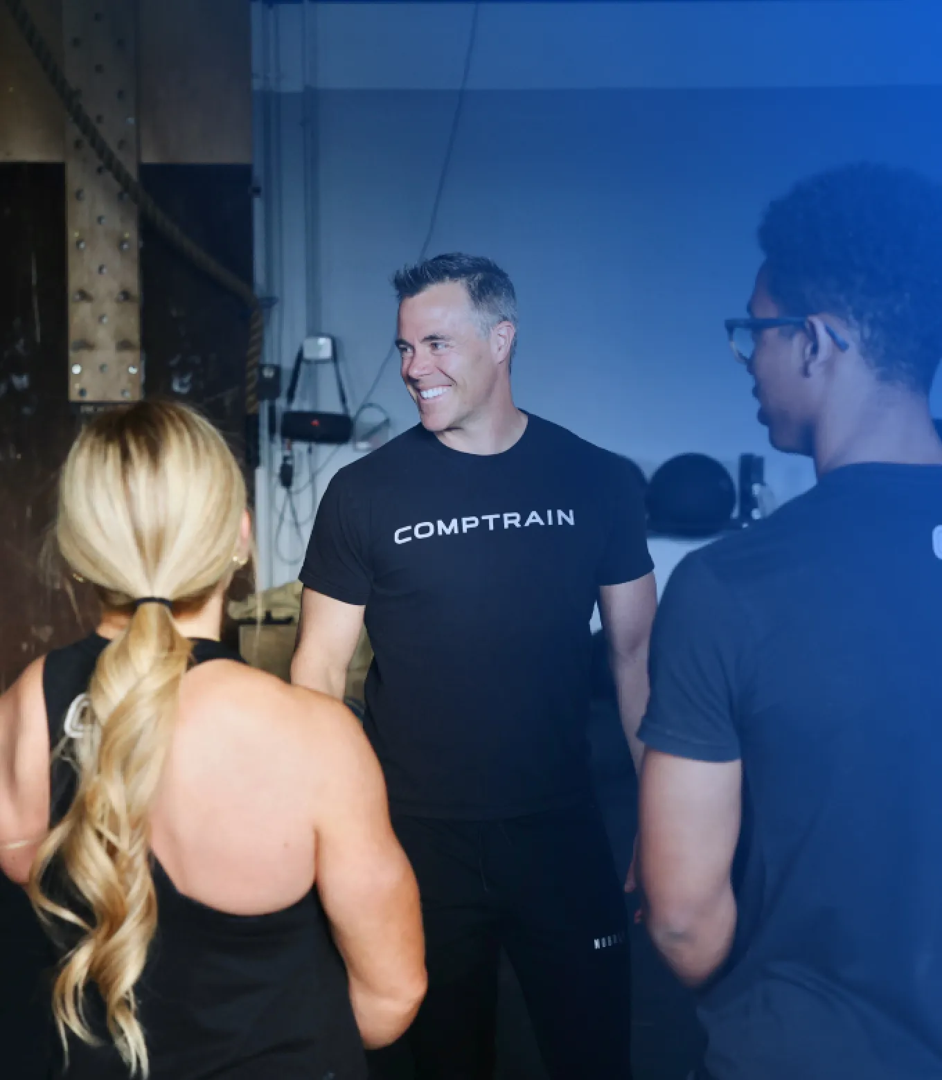 Three people in athletic wear having a conversation, with one man smiling and wearing a black COMPTRAIN shirt.