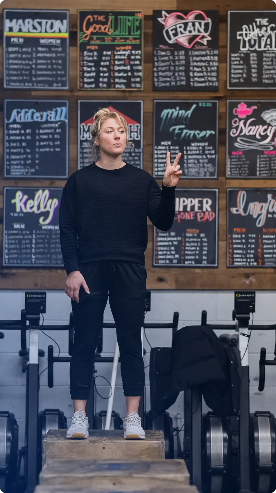 Woman standing on a wooden box in a gym, holding up two fingers in front of a wall with various workout leaderboards.