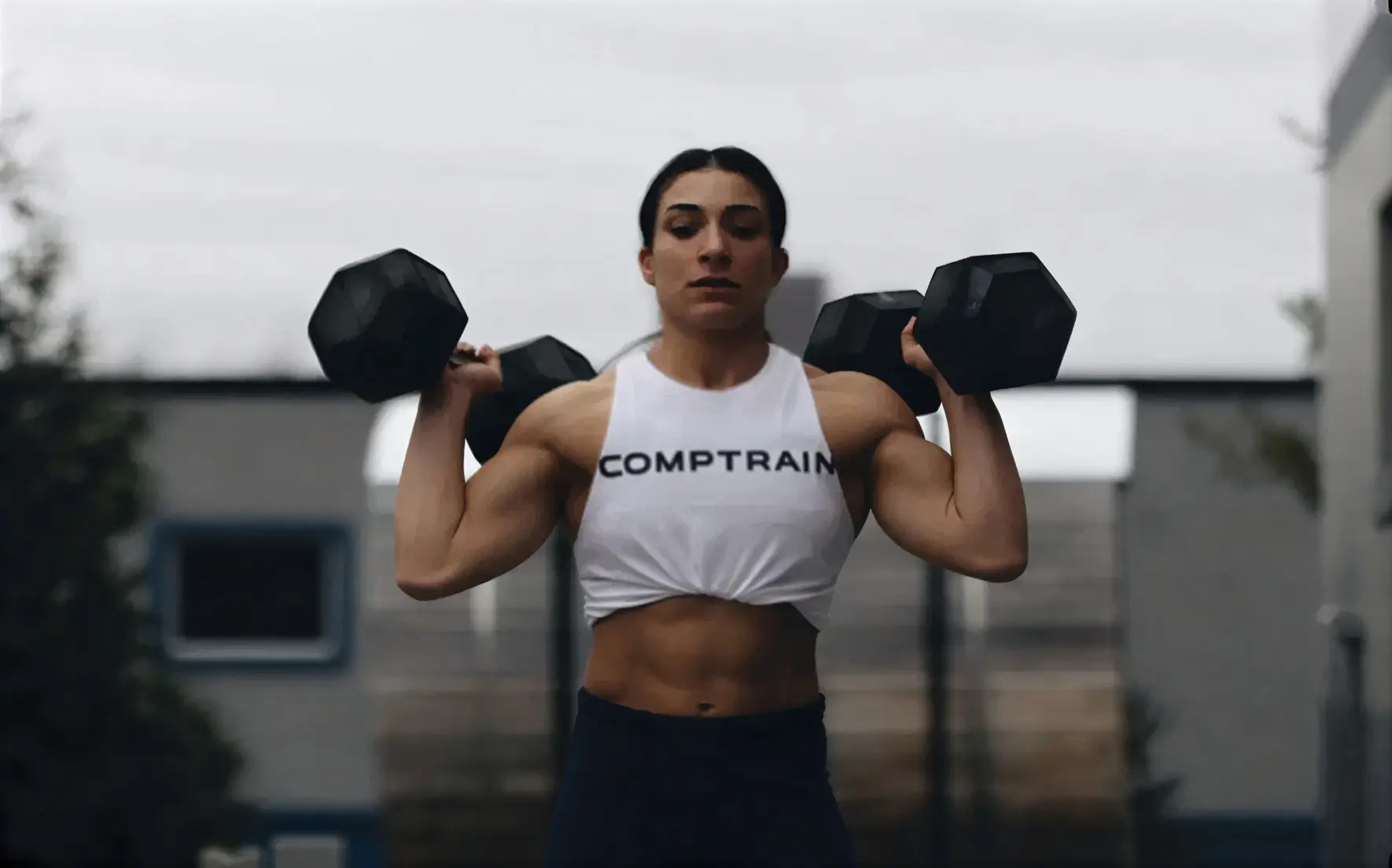 Athletic woman lifting dumbbells outdoors wearing a white tank top with the text 'COMPTRAIN'.