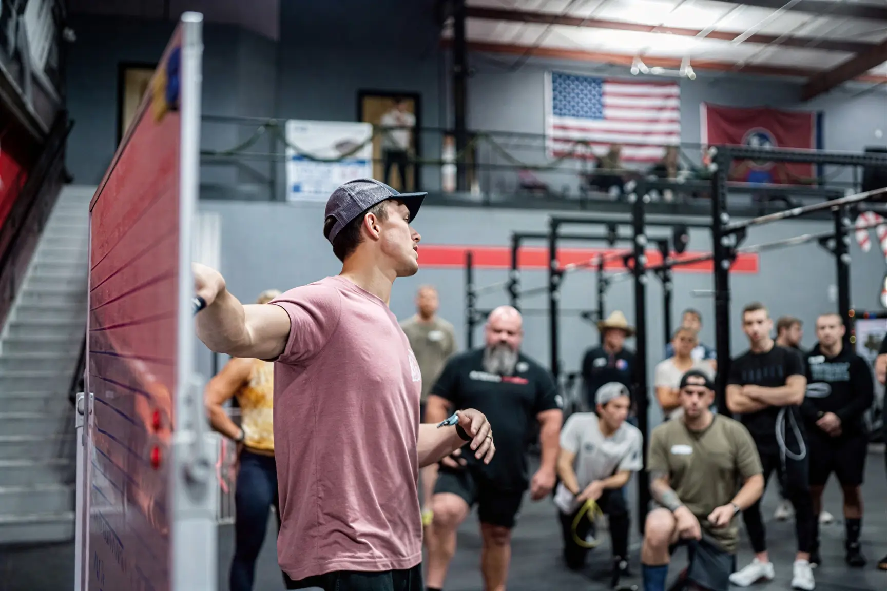 Man in a pink shirt and cap speaking to a group of people in a gym with American and Tennessee flags in the background.