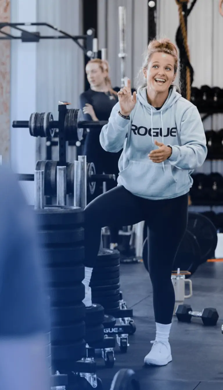 Smiling woman in a gym wearing a light gray hoodie with 'ROGUE' printed on it, lifting her knee and gesturing with her hands.