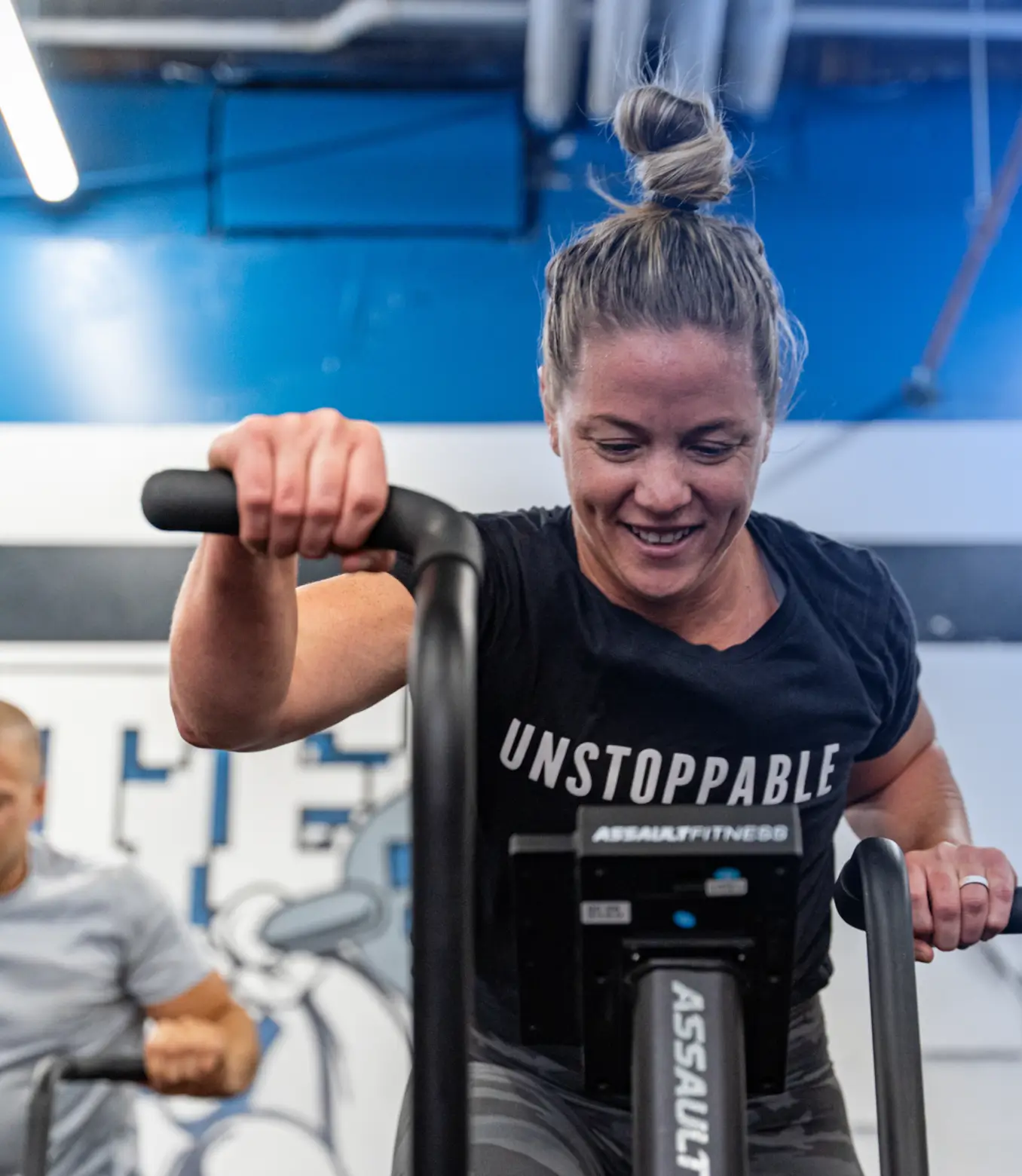 Smiling woman in an 'UNSTOPPABLE' shirt working out on an Assault Fitness exercise bike in a gym.
