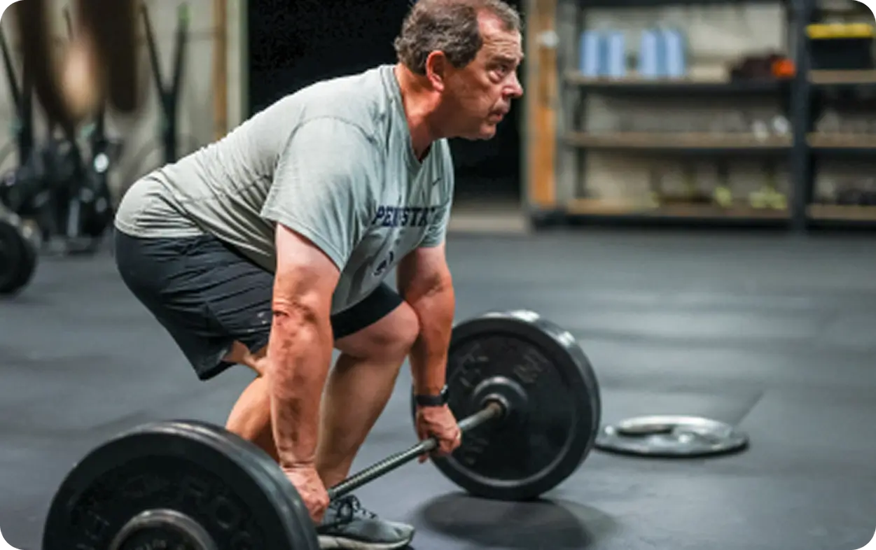Middle-aged man in gray shirt and black shorts lifting a barbell in a gym.