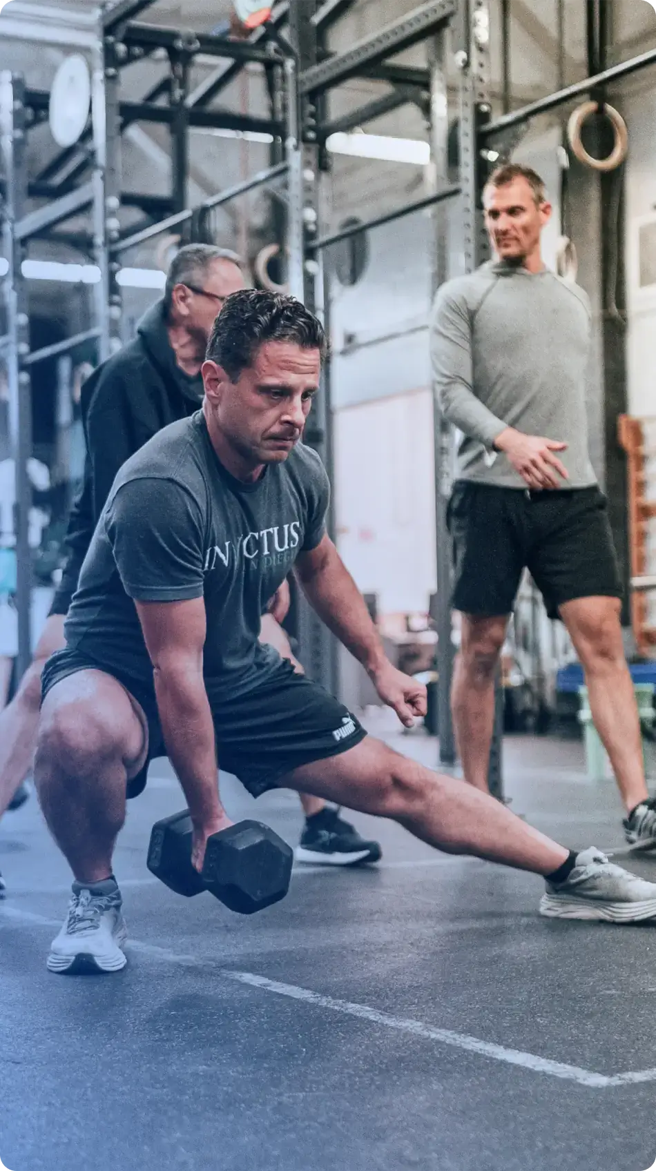 Man performing a side lunge with a dumbbell in a gym while two men watch.