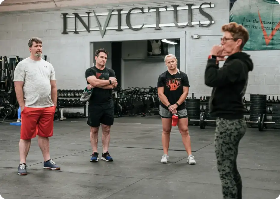 Four people in workout clothes standing and listening inside a gym with 'INVICTUS' sign on the wall.