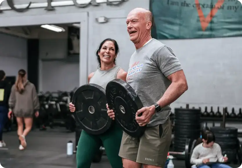 Two smiling adults lifting black weight plates inside a gym with people in the background.