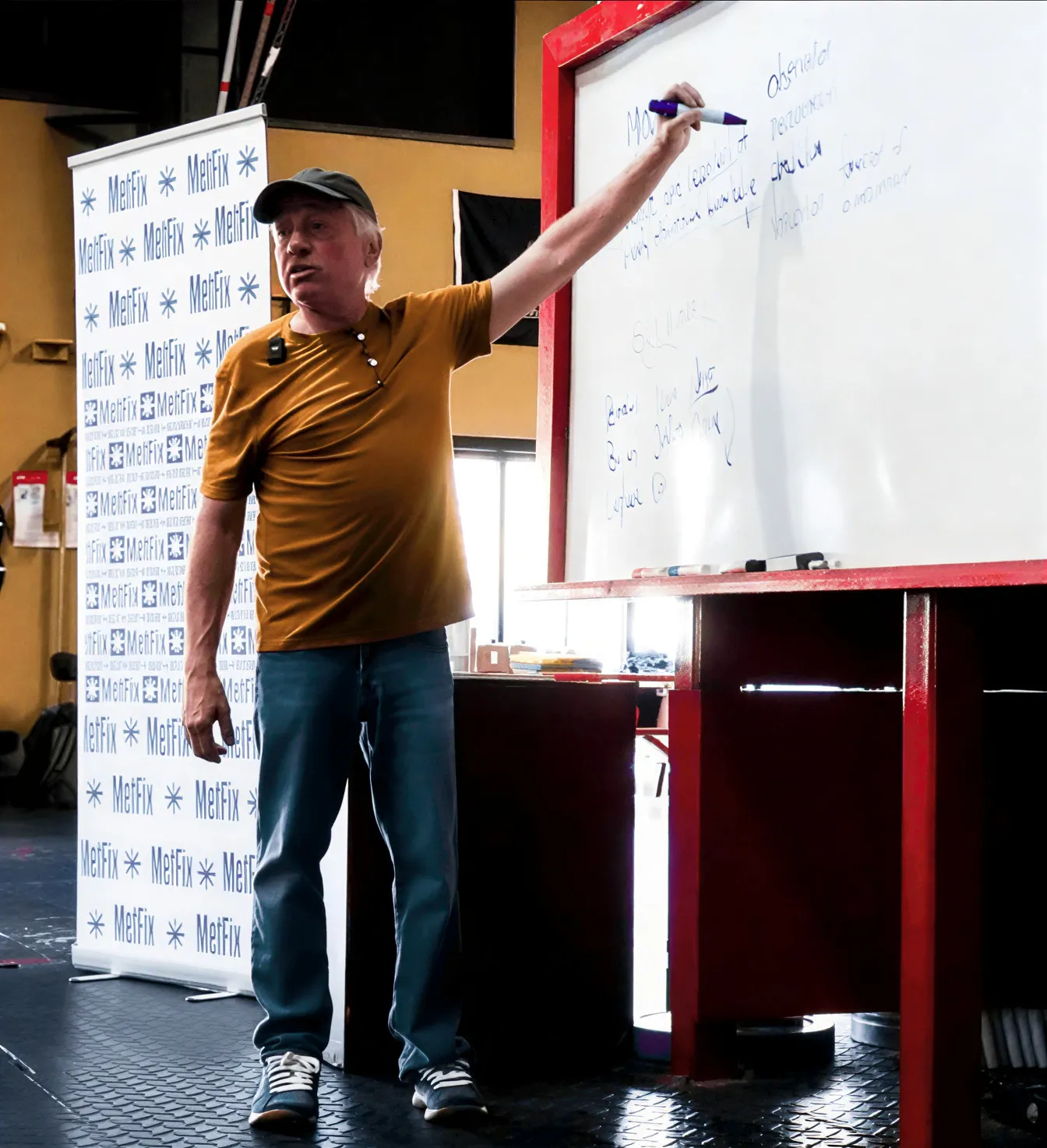 Man in a brown shirt and cap pointing at notes on a whiteboard in a classroom or workshop setting.