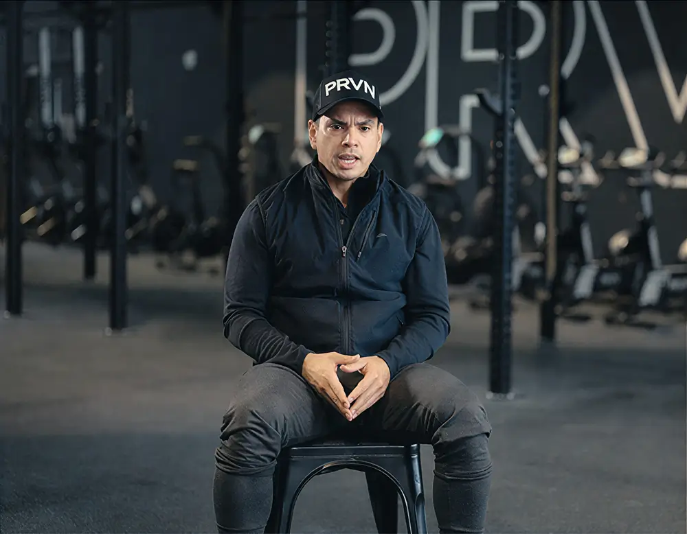 Man wearing a PRVN cap and black athletic clothing sitting on a stool in a gym with workout equipment in the background.