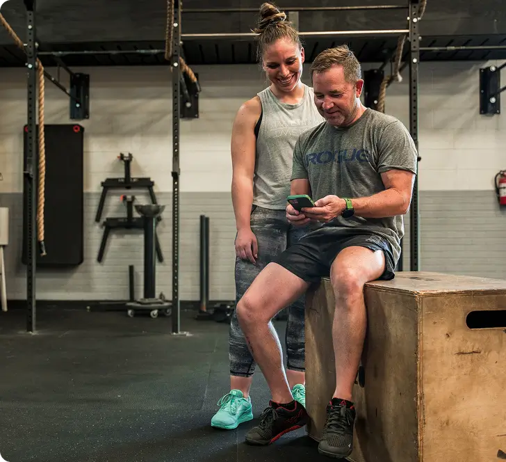 Man sitting on a wooden box in a gym looking at his phone while a woman in workout clothes stands beside him smiling.