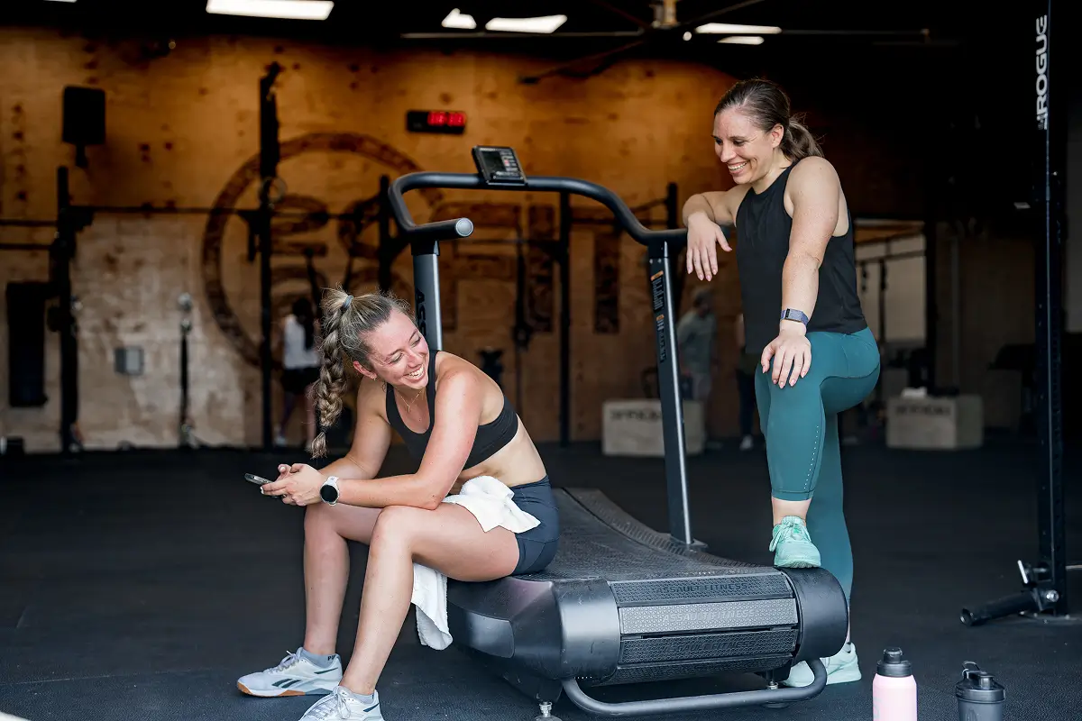 Two women in athletic wear smiling and chatting next to a treadmill in a gym.
