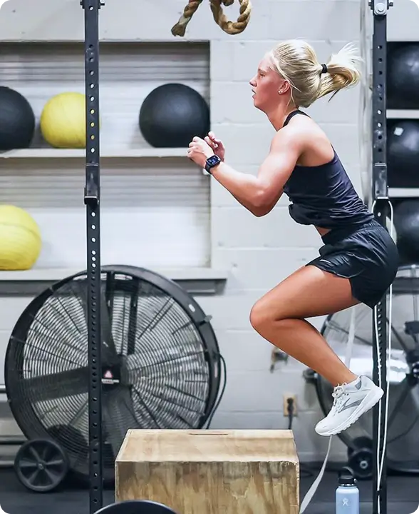 Athletic woman performing a box jump exercise in a gym with fitness balls and a large fan in the background.