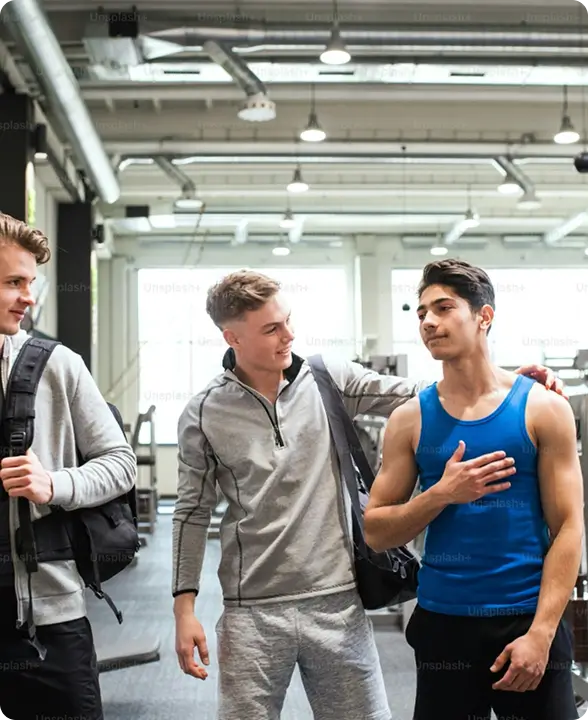 Three young men in a gym, one in a blue tank top with hand on chest, another in gray athletic wear smiling and patting his shoulder, and a third man holding a backpack strap.