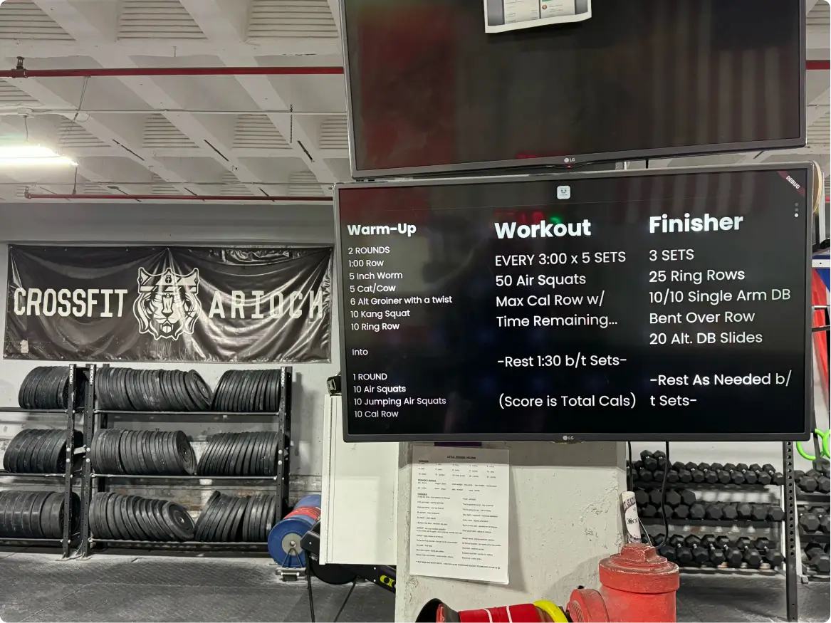 CrossFit gym interior with tire racks and two large screens displaying warm-up, workout, and finisher routines.