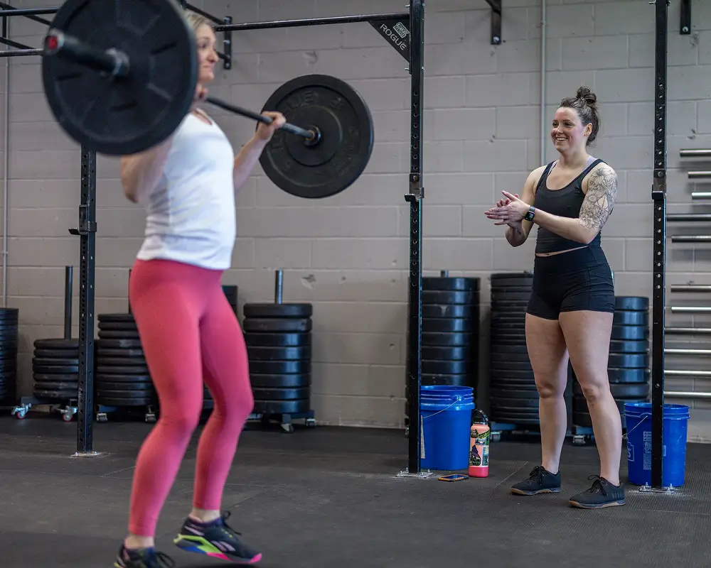 Woman in pink leggings lifting a barbell while another woman in black shorts claps encouragement in a gym.