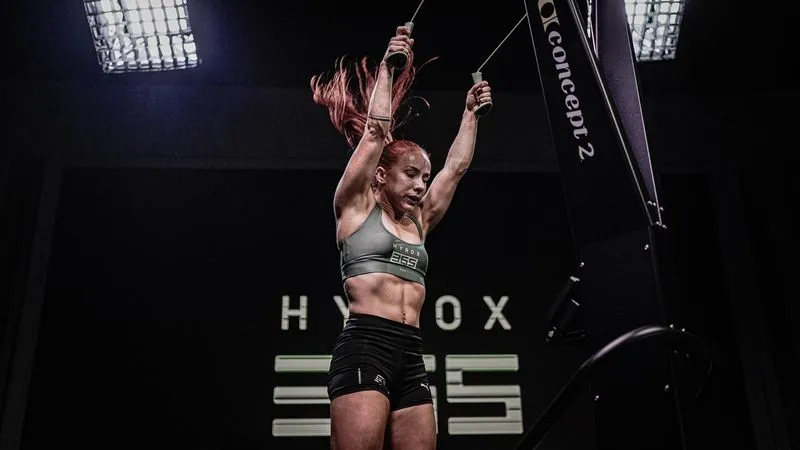 Athletic woman with red hair using a Concept2 ski ergometer in a gym with black walls and bright ceiling lights.