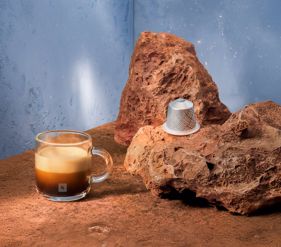 Glass cup of espresso with foam and a decorated coffee capsule on reddish rocks and surface.