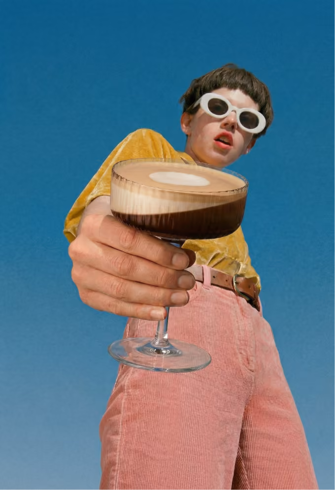 Person in white sunglasses holding a glass of frothy coffee against a clear blue sky.