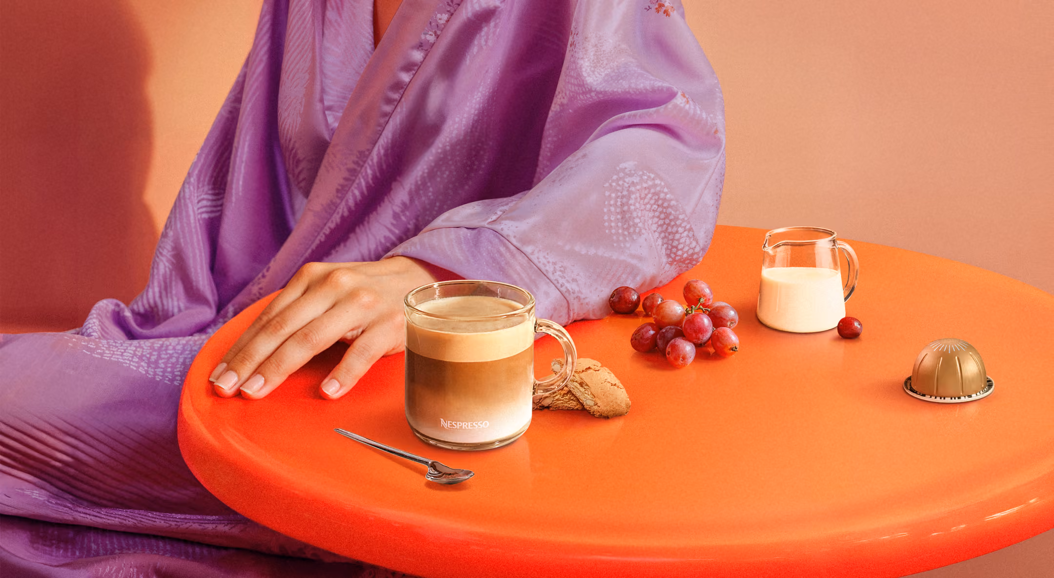 Person in purple robe at orange table with coffee, grapes, milk jug, cookie, and coffee capsule.