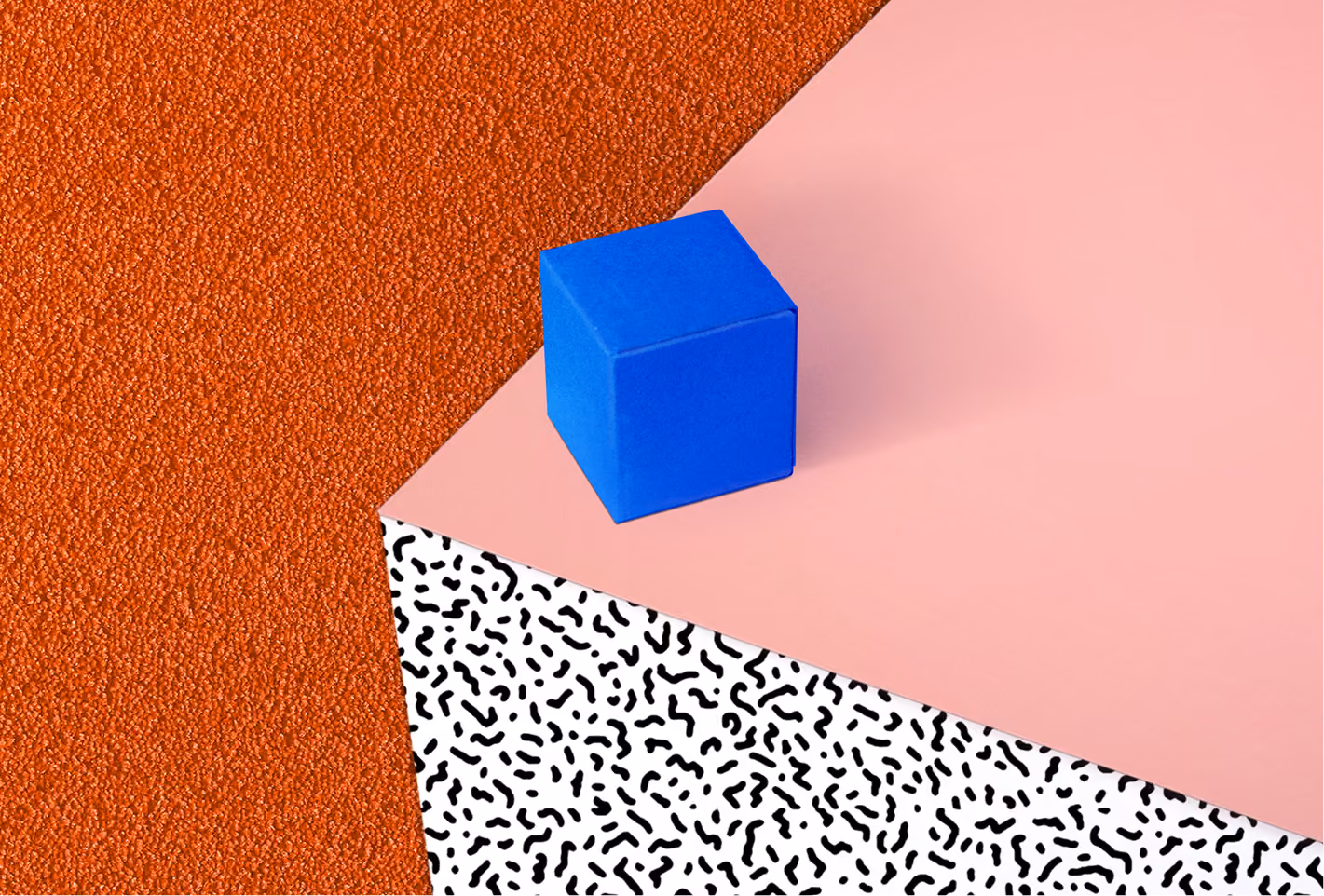 Blue cube on a pink surface above a white pattern with black squiggles and an orange textured background.