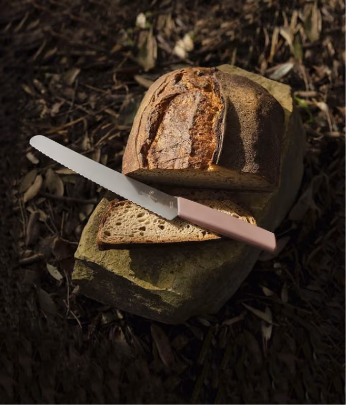 Sliced loaf of rustic bread on a rock with a serrated knife resting on a slice outdoors.
