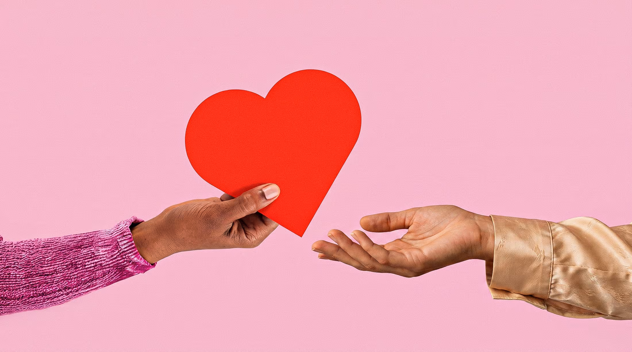 A hand in a pink sweater holds a red heart toward an open hand in a beige shirt on a pink background.