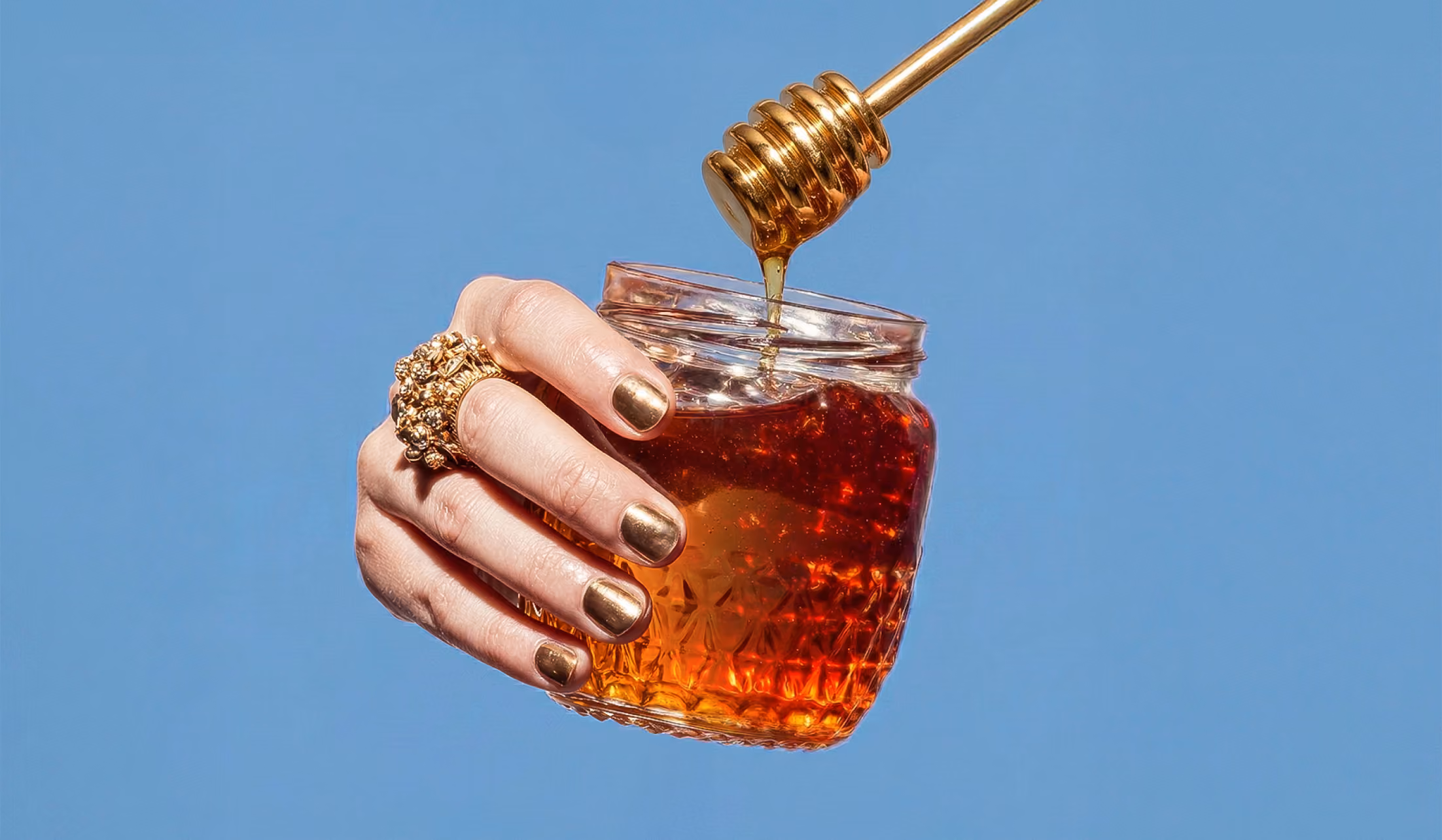 Hand with gold nail polish and ring holding a jar of honey while honey drips from a honey dipper.